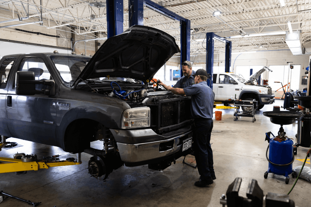 Diesel truck noises, diesel repair in Littleton, CO by Branch Automotive. Image of two technicians servicing a lifted Ford Super Duty with the hood open, performing diagnostics and maintenance inside a professional diesel repair shop.